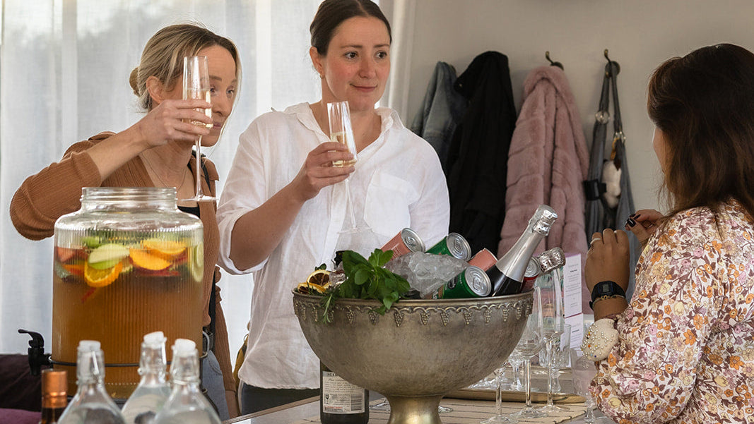 Three women standing behind a wooden bar counter in a bright indoor setting, holding glasses and preparing drinks. A large glass beverage dispenser with orange slices, several clear bottles, a metal bowl of ice and greenery, and champagne bottles are arranged on the counter.