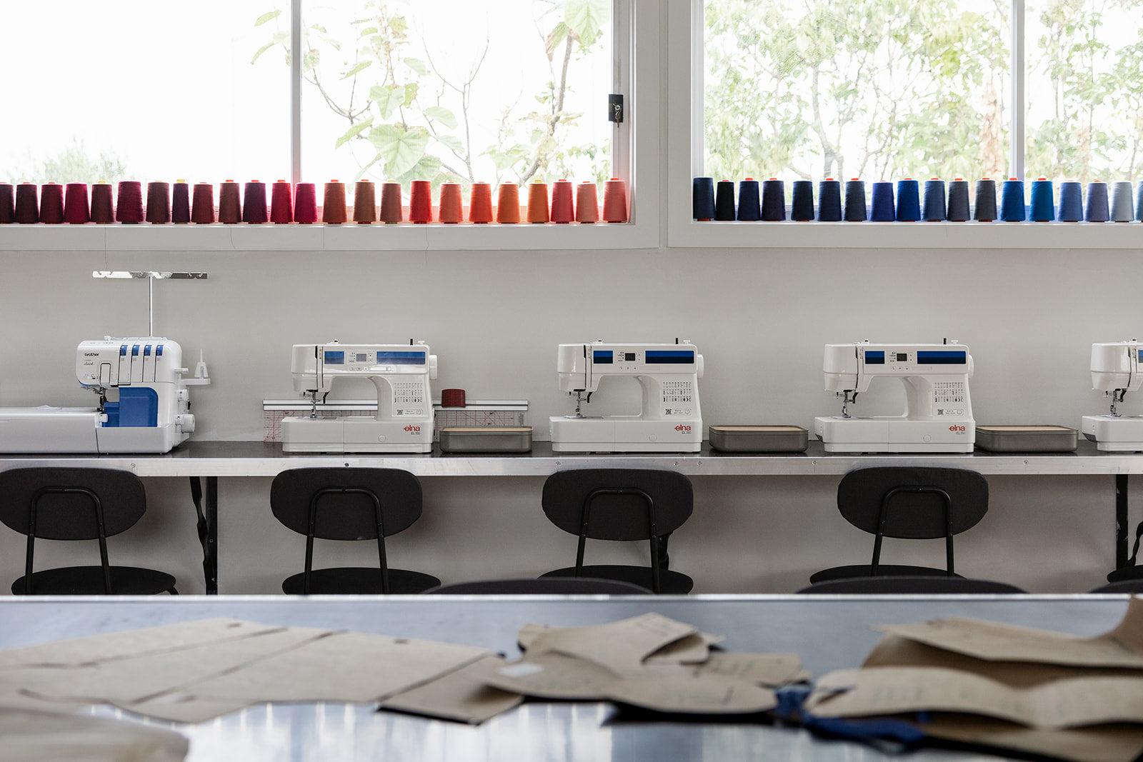 Row of sewing machines on a table with fabric in front, against a windowed wall.