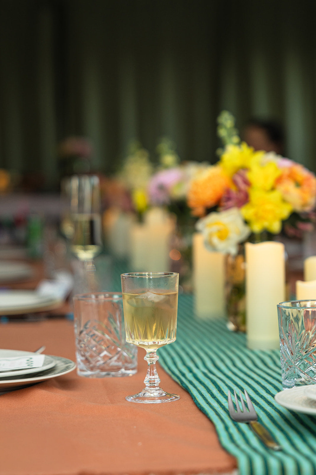 A styled dining table setting with a green striped runner, terracotta tablecloth, clear glassware, and a blurred floral centerpiece in the background.