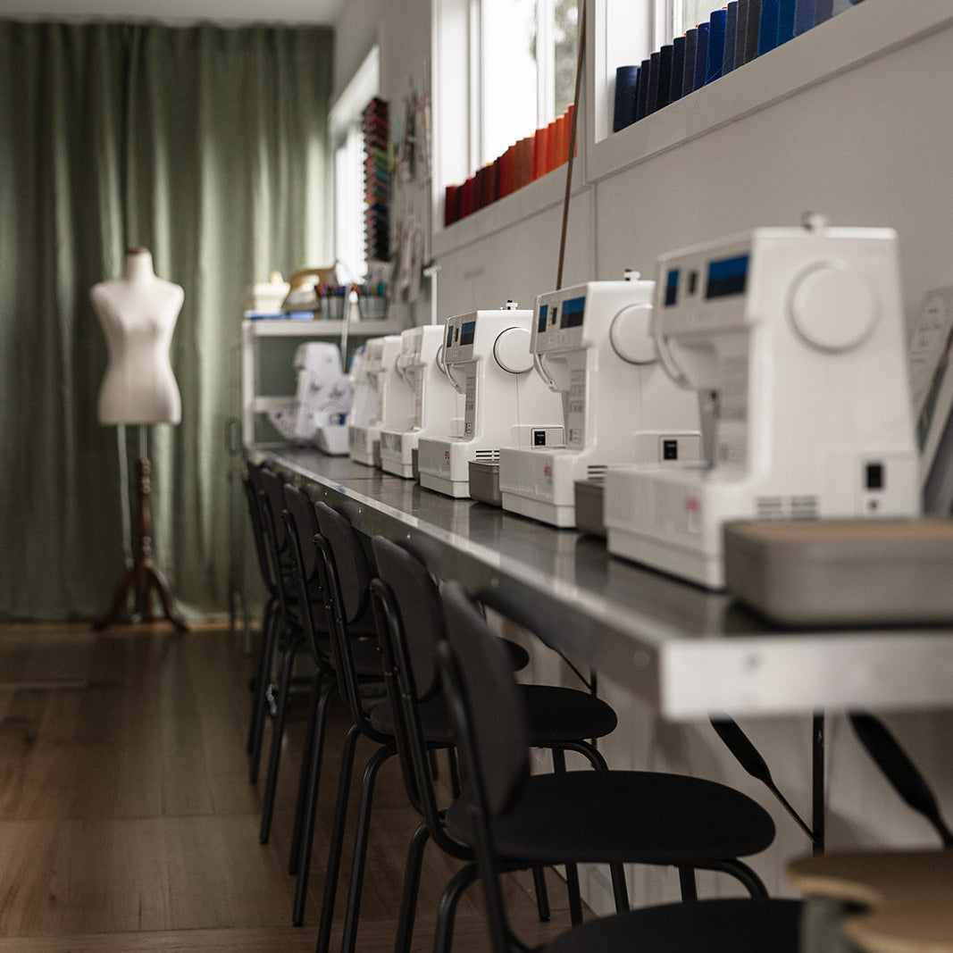Row of sewing machines on a table in a room with chairs and a mannequin.