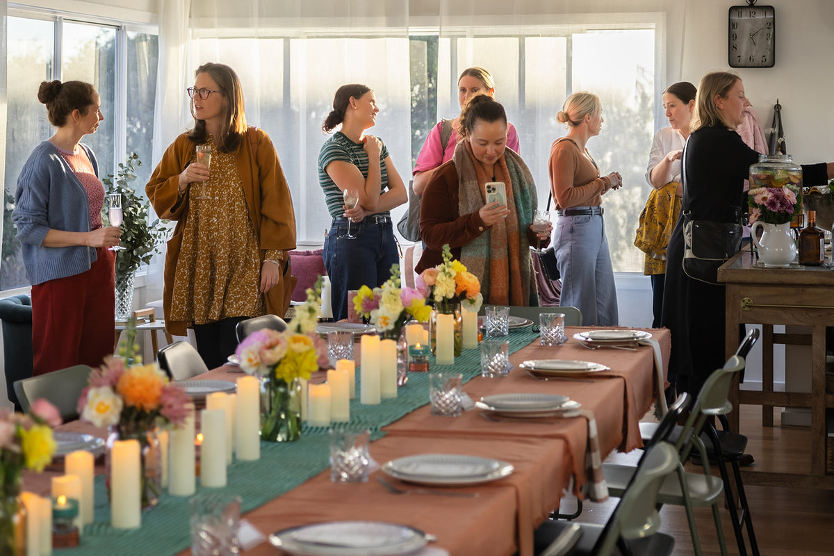 A group of women stand around a decorated event table with candles, flowers, plates, and chairs in a bright indoor space with large windows.